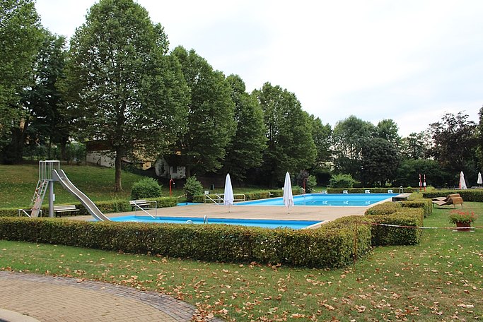 Das Bild zeigt ein Schwimmbad. Im Vordergrund befindet sich eine Wiese und eine Hecke. Links sieht man eine Rutsche, die in ein Schwimmbecken führt. In der Mitte befindet sich das blaue Schwimmbecken. Auf den Seiten sieht man eine Liegewiese mit Bäumen und eingeklappten weißen Sonnenschirmen.