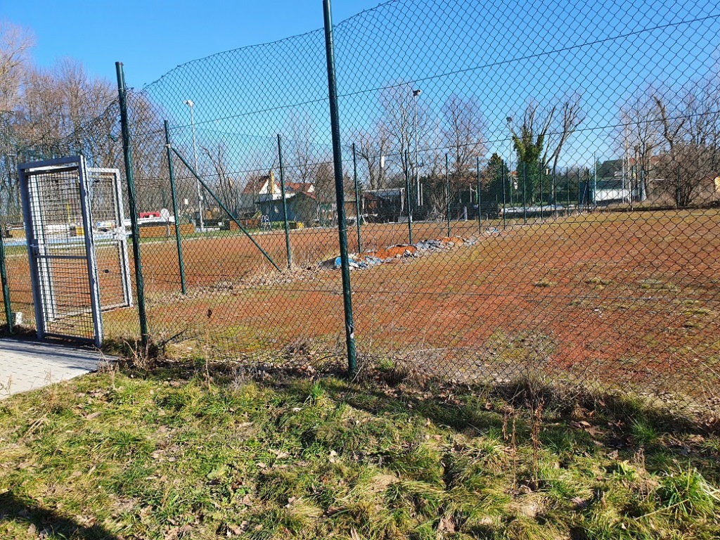 Das Bild zeigt einen hohen Maschendrahtzaun, welcher einen grasbewachsenen Tennisplatz umzäunt.