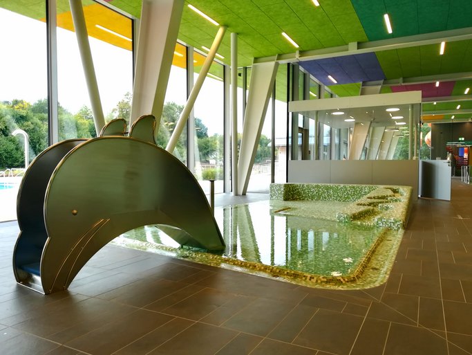 Das Bild zeigt ein Kinderbecken mit Delfinrutsche in einem bunte gestalteten Hallenbad.