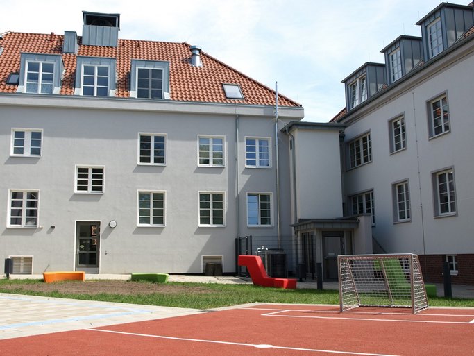 Das Bild zeigt einen Innenhof mit rotem Sportfeld und Tor, Rasen- und Pflasterfläche sowie Sitzgelegenheiten, umrahmt von grauen Gebäuden.