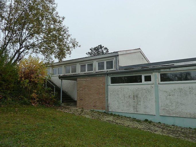 Das Bild zeigt das zu renovierende Gebäude von außen, welches in sichtlich schlechtem Zustand ist. Am linken Rand steht ein Baum. Im Vordergrund befindet sich eine Wiese.