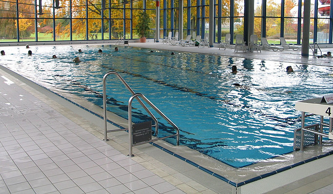 Das Bild zeigt ein Schwimmbecken in einem Hallenbad mit ein paar Personen darin.