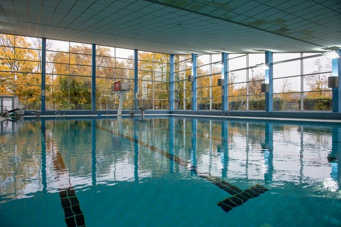 Das Bild zeigt ein Schwimmbecken mit Sprungturm in einem Hallenbad, im Hintergrund ist eine großflächige Glasfassade zu sehen.