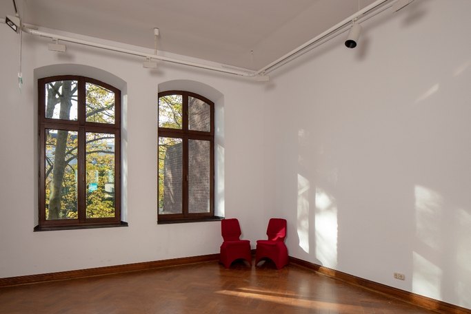 Das Bild zeigt einen lichtdurchfluteten leeren Innenraum mit zwei Fenstern und zwei rot bezogenen Stühlen in der Ecke.