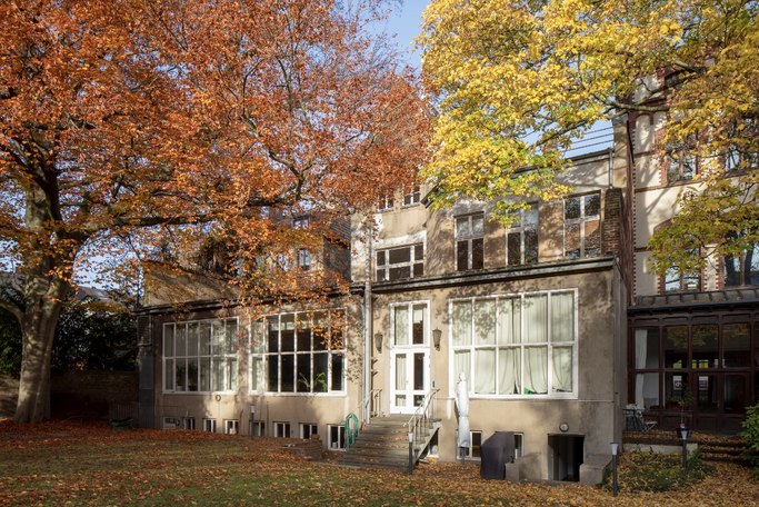 Das Bild zeigt ein braunes Gebäude mit Fensterfront von außen, umgeben von herbstlichen Laubbäumen.