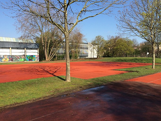 Das Foto zeigt einen roten Sportplatz. Grüne Flächen mit Bäumen ergänzen das Sportfeld. Im Hintergrund sieht man das Sportgebäude.