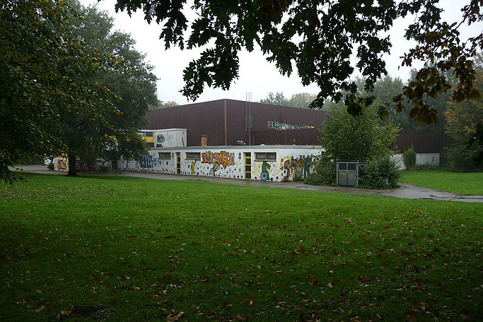 Das Bild zeigt ein braunes Flachdachgebäude mit weißen Vorbau, gelben Fenster und Graffiti an den Wänden, umgeben von Grün.