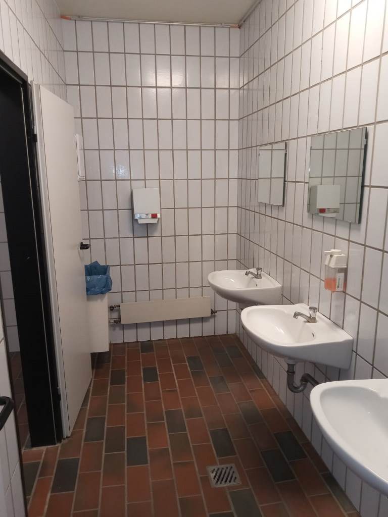 Man sieht eine Toilette mit drei Waschbecken und Spiegeln darüber auf der rechten Wandseite.