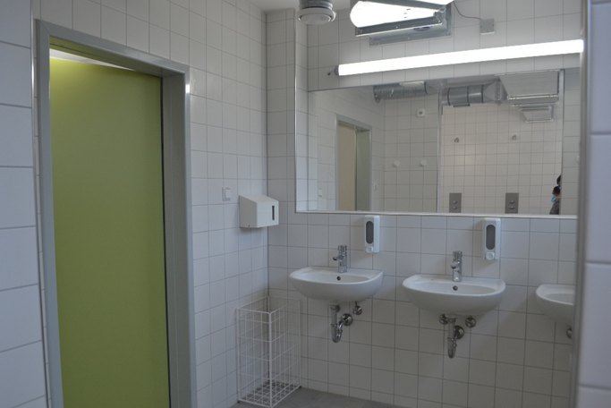 Das Bild zeigt einen weiß-gefliesten Sanitärbereich, bei dem mehrere Waschbecken und Spiegel in einer Reihe sind.
