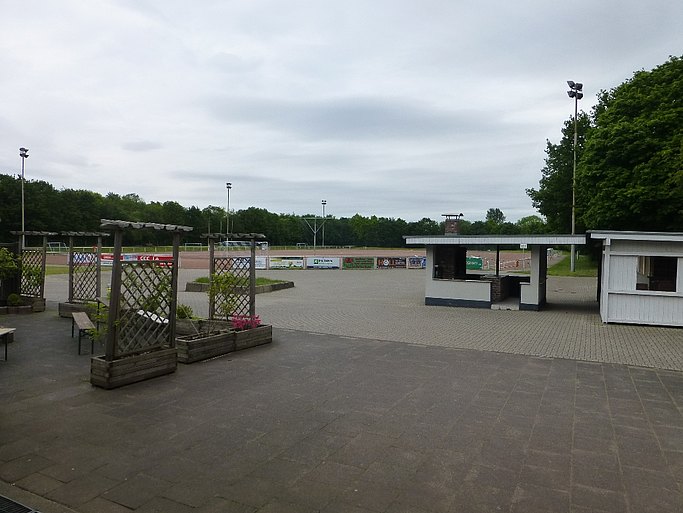 Man sieht die Sportanlage vor der Sanierung. Auf der rechten Seite befinden sich zwei weiße Kioske, links mehrere Bänke und Rankgitter. Im Hintergrund sieht man das Sportfeld.