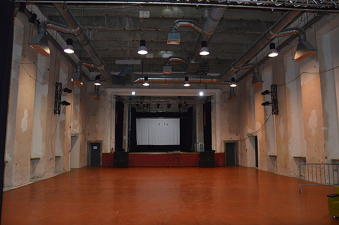 Das Bild zeigt einen leeren Saal mit Bühne und ohne Fenster.