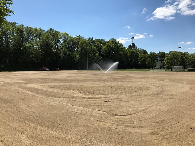 Das Bild zeigt die Sanierung des Rasenplatzes in vollem Gange. Die Rasenfläche ist abgetragen und neu eingesäht. Ein Rasensprenkler steht in der Mitte. 