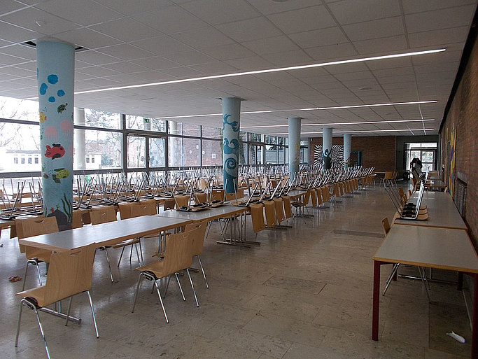 Das Bild zeigt einen großen Raum mit Tischreihen und Stühlen, durchzogen von bemalten Säulen, im Hintergrund ist eine Fensterfront zu sehen.