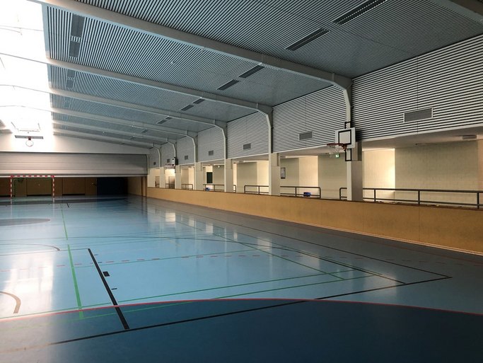 Das Bild zeigt eine Sporthalle mit blauem Boden, bunten Linien und Basketballkorb.