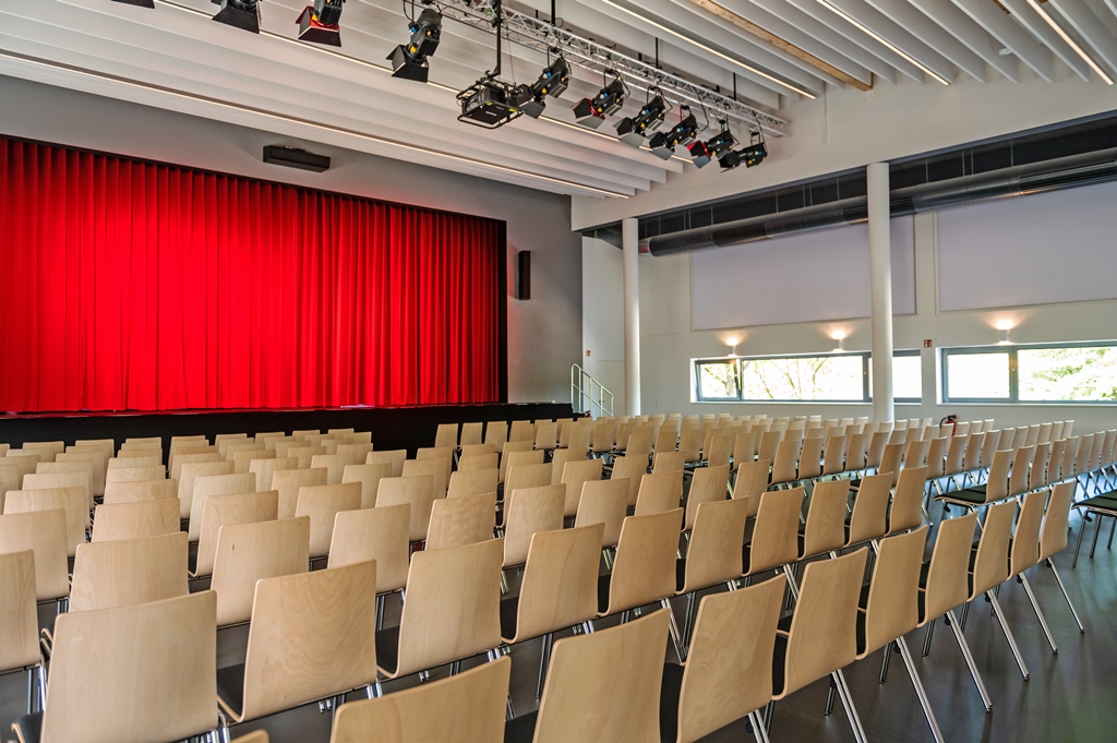 Das Bild zeigt eine bestuhlte Halle mit Bühne und rotem Vorhang.