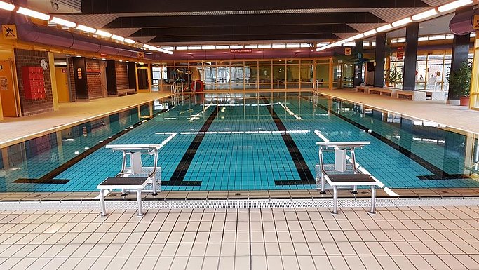 Das Bild zeigt ein Schwimmbecken in einem Hallenbad, im Vordergrund sind Startblöcke zu sehen.