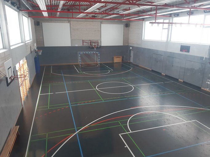 Das Bild zeigt eine Sporthalle mit grauem Boden und bunten Markierungen sowie diversen Sportgeräten.