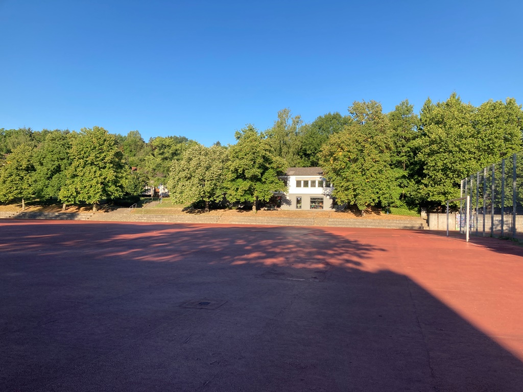 Das Foto zeigt eine Sportanlage mit rotem Boden, umgeben von Bäumen.