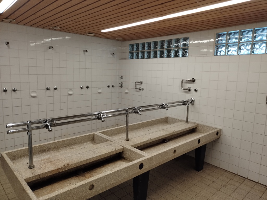 Das Bild zeigt Sanitäranlagen, im Vordergrund sind Waschbecken zu sehen, im Hintergrund mehrere Duschen.