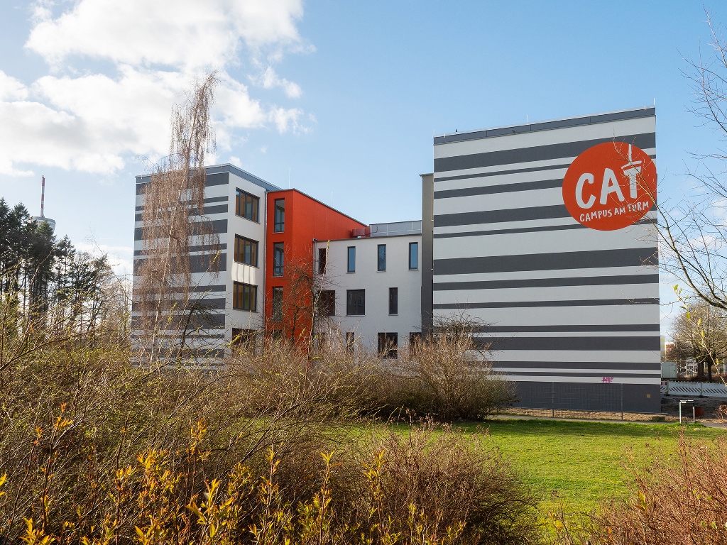 Das Bild zeigt ein grau-oranges Gebäude mit der Aufschrift CAT Campus am Turm, im Vordergrund sind Grünflächen und Sträucher zu sehen.