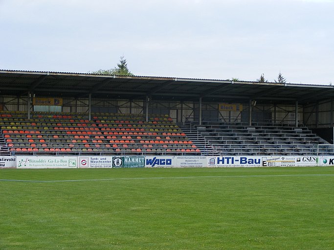 Das Bild zeigt eine Zuschauertribüne, im Vordergrund ist ein Fußballplatz zu sehen.