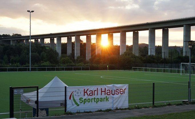 Das Bild zeigt einen Fußballplatz bei Sonnenuntergang, im Hintergrund ist eine Brücke zu sehen.