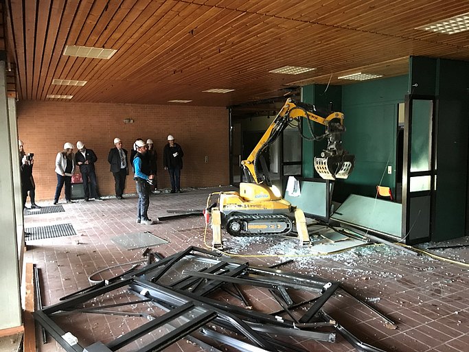 In dem Bild stehen mehrere Personen mit Helm. Ein kleiner Bagger reißt einen Raum ein. Am Boden liegen Rahmen.