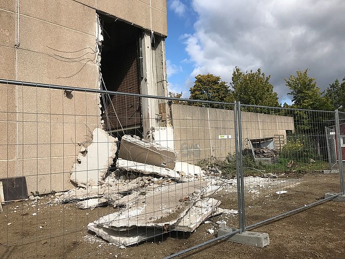 Das Bild zeigt ein Gebäude, von dessen Fassade ein Teil abgerissen wurde. Im Vordergrund steht ein Bauzaun.