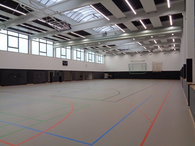 Das Bild zeigt eine hell gestaltete Sporthalle mit großzügigen Fenstern, hellgrauem Boden und verschiedenen Sportgeräten.