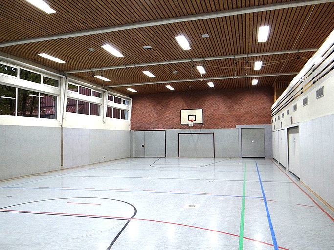 Das Bild zeigt eine Sporthalle mit hellem Boden, bunten Markierungen und Basketballkorb.