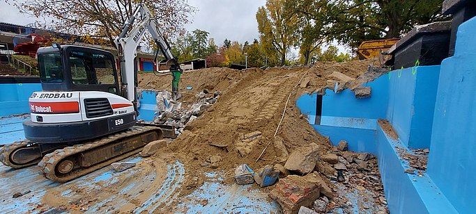 Man sieht Bauarbeiten in einem Freibad. Der Bagger steht im leeren Schwimmbecen und entfernt die Stirnseite.