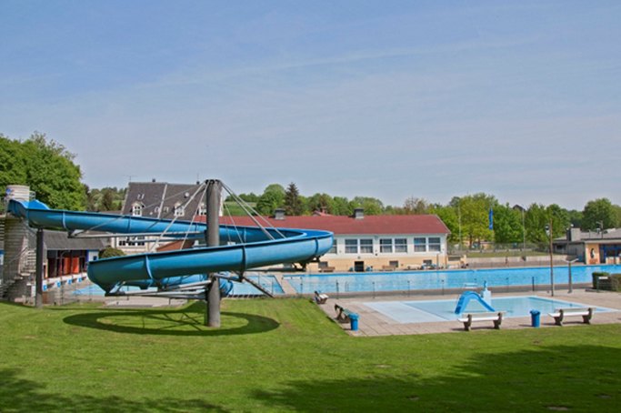 Das Bild zeigt Schwimmbecken und Wasserrutsche in einem Freibad, im Hintergrund ist ein Gebäude zu sehen.