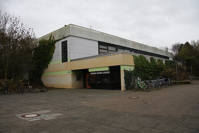Das Bild zeigt den Eingangsbereich zu einem verwitterten Gebäude mit Flachdach, im Vordergrund ist sind eine Pflasterfläche und Fahrradständer zu sehen.