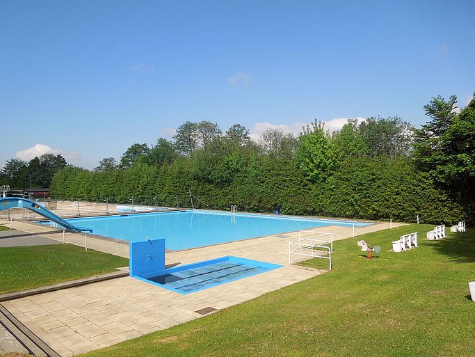 Das Bild zeigt ein Schwimmbecken, in welches eine Rutsche vom linken Rand führt. Im Vordergrund sieht man eine Liegewiese. Die Beckenumrandung ist gepflastert.