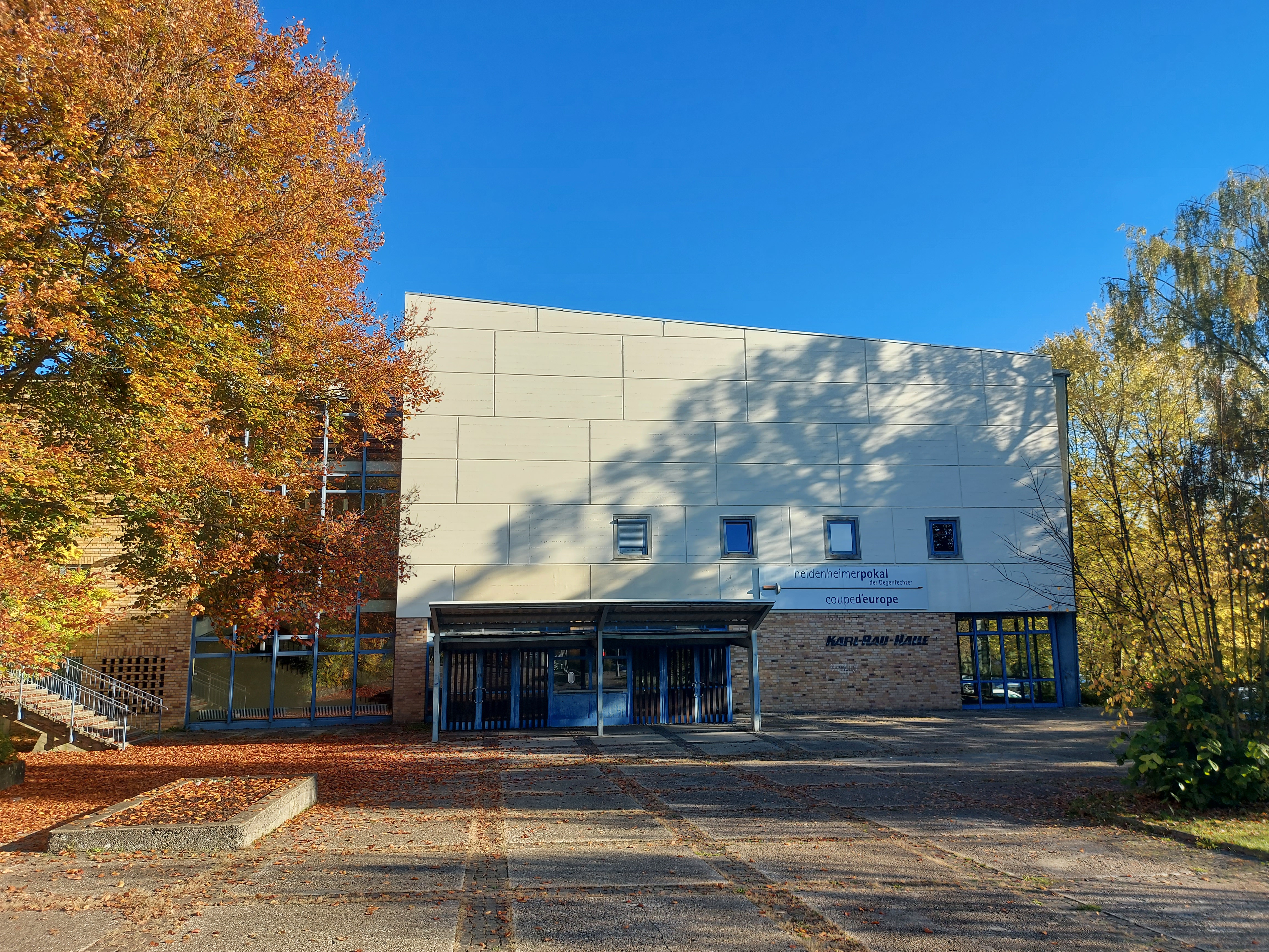 Das Bild zeigt die Sporthalle von vorne. Das Dach verläuft schräg nach unten und der Eingang ist überdacht. Neben dem Vorplatz stehen rechts und links Bäume.