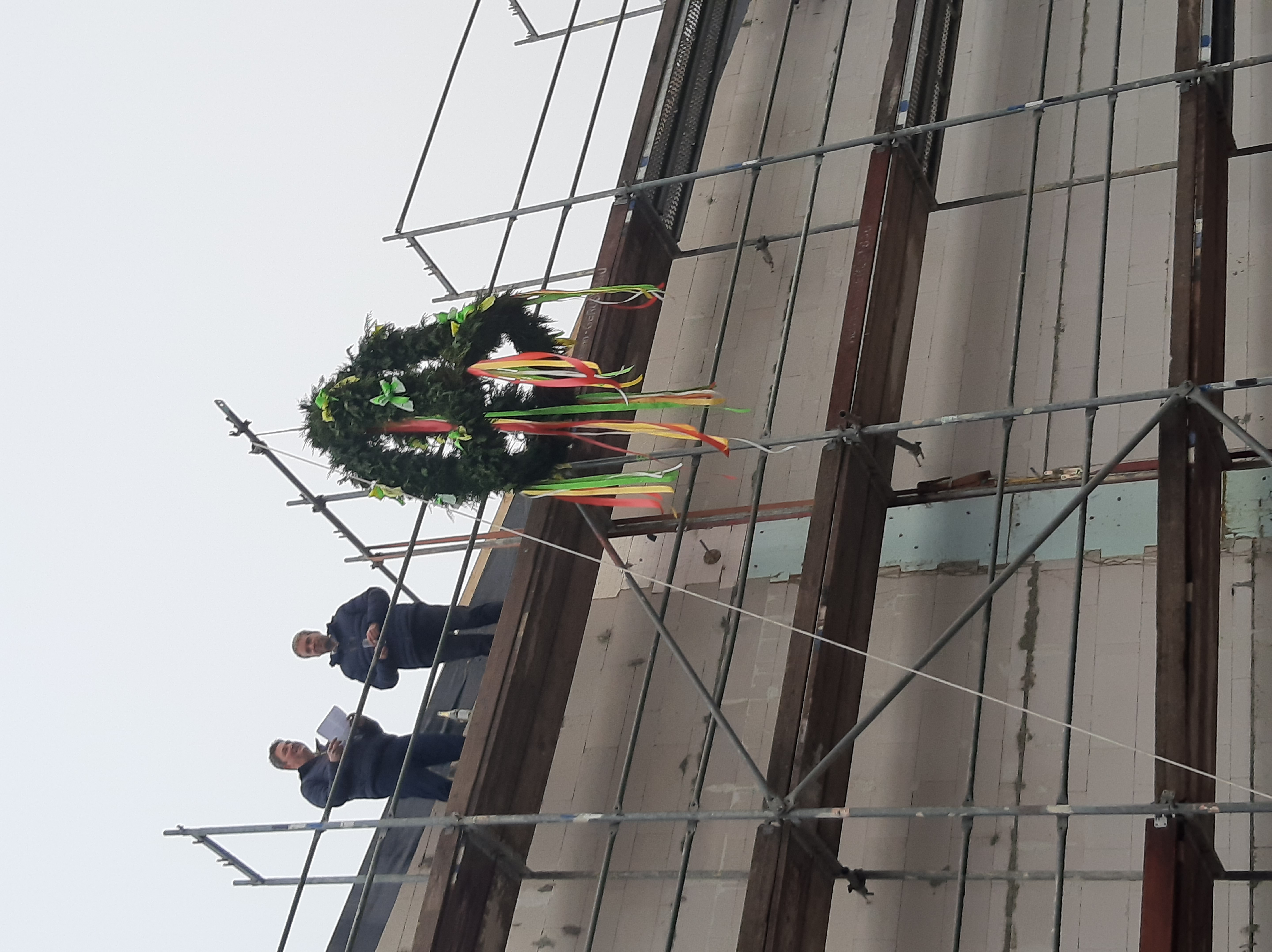 Das Bild zeigt ein Gebäude, das von einem Gerüst eingezäunt ist. Auf dem Dach stehen zwei Männer. Ein Richtkranz mit Bändern hängt an der Einzäunung.