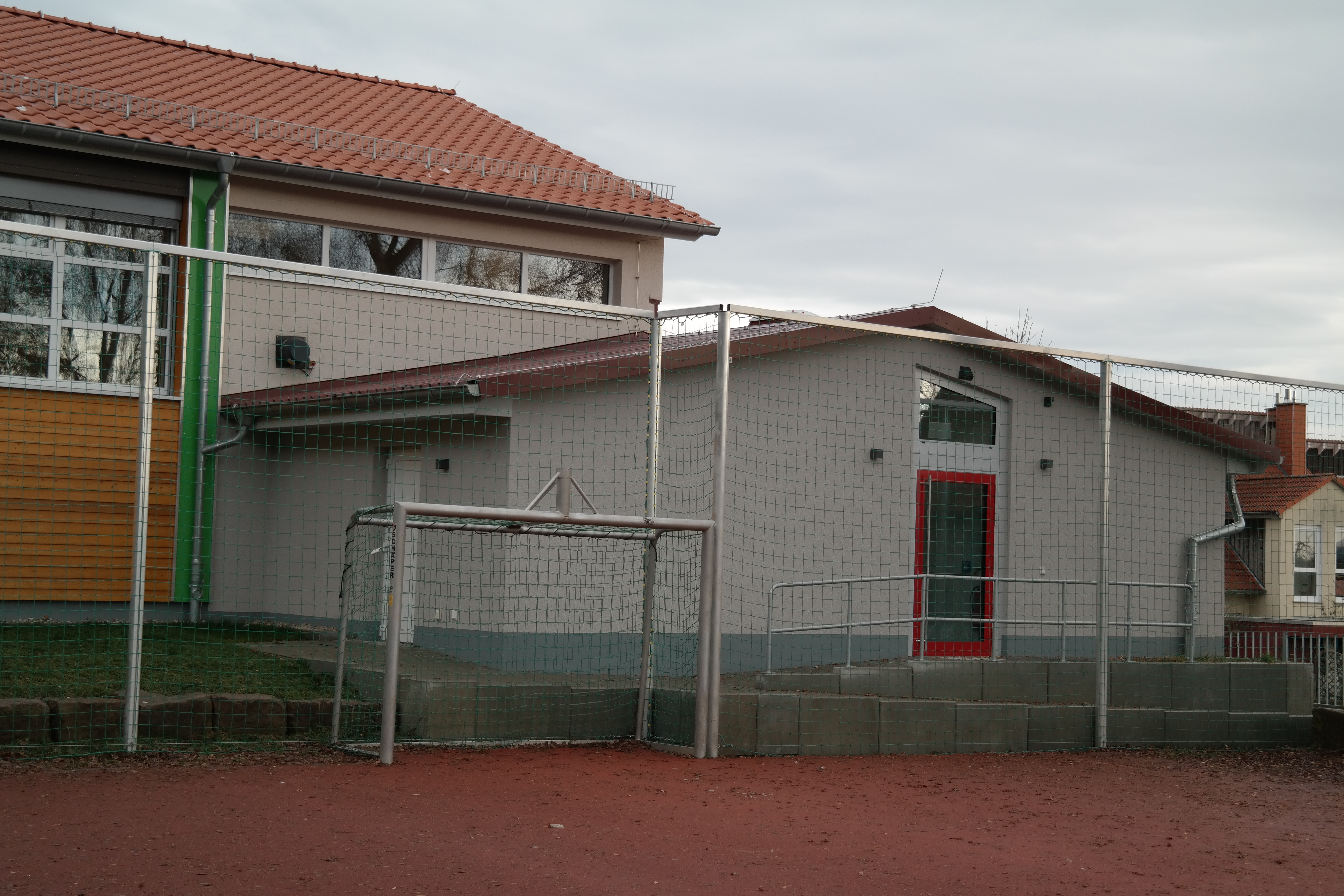 Das Bild zeigt die Rückseite einer Sporthalle. Im Vordergrund sieht man einen Tennenplatz mit Tor und Fangzaun.