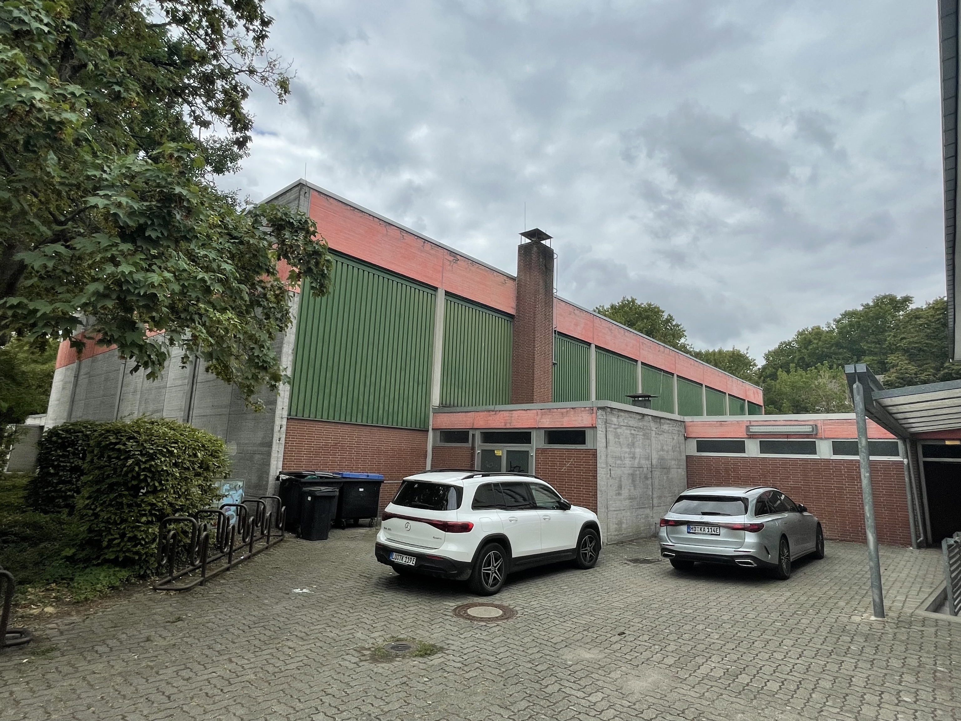 Quelle: S. Nuccio Das Bild zeigt eine Nord-Ost Ansicht einer sanierungsbedürftigen Halle. Man erkennt im Vordergrund zwei Autos.