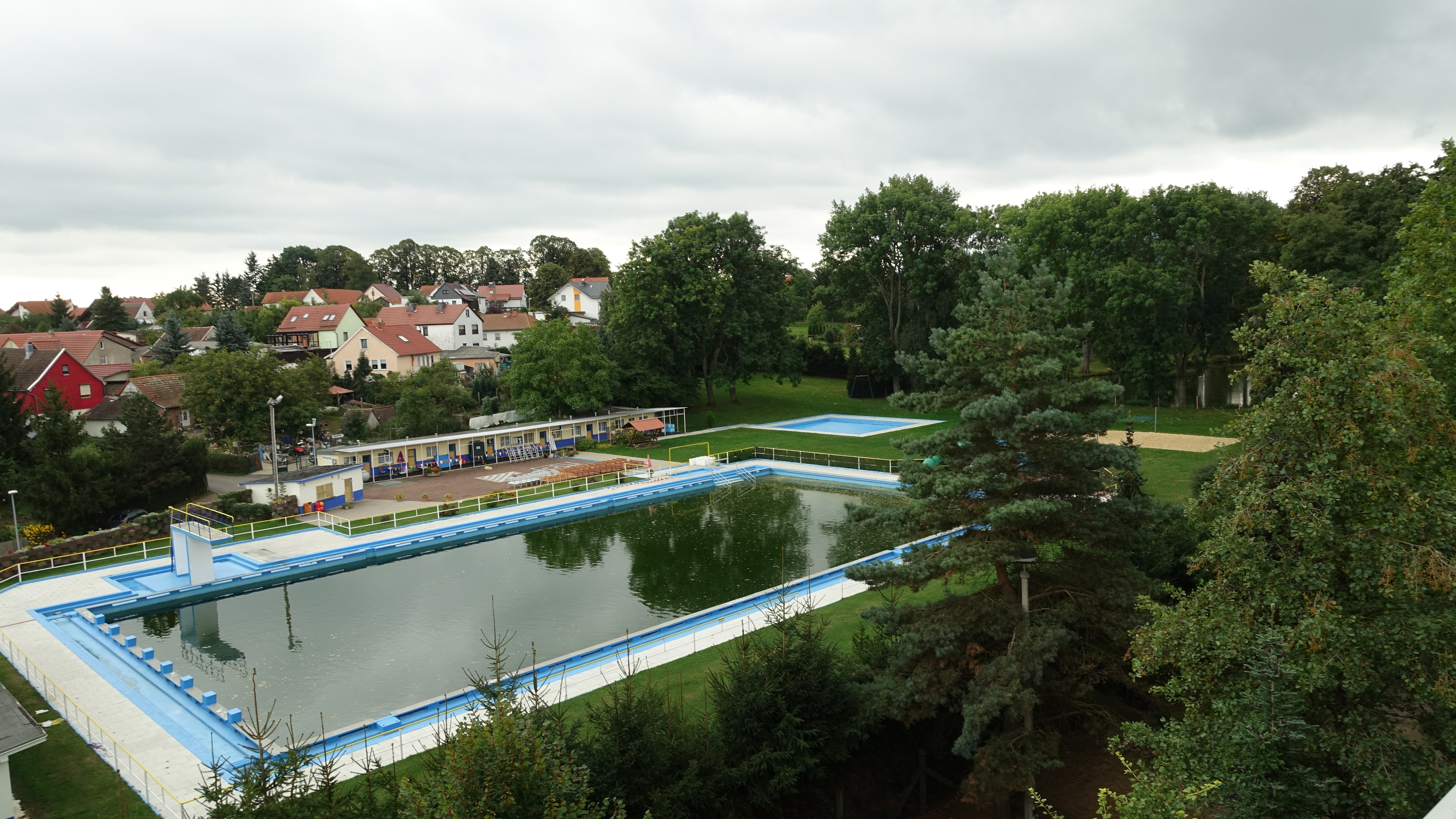 Das Bild zeigt ein Freibad von oben. Das Wasser im Schwimmerbecken ist dunkelgrün. Rundherum befinden sich Liegewiesen, ein Kinderbecken und ein Beachvolleyballplatz.