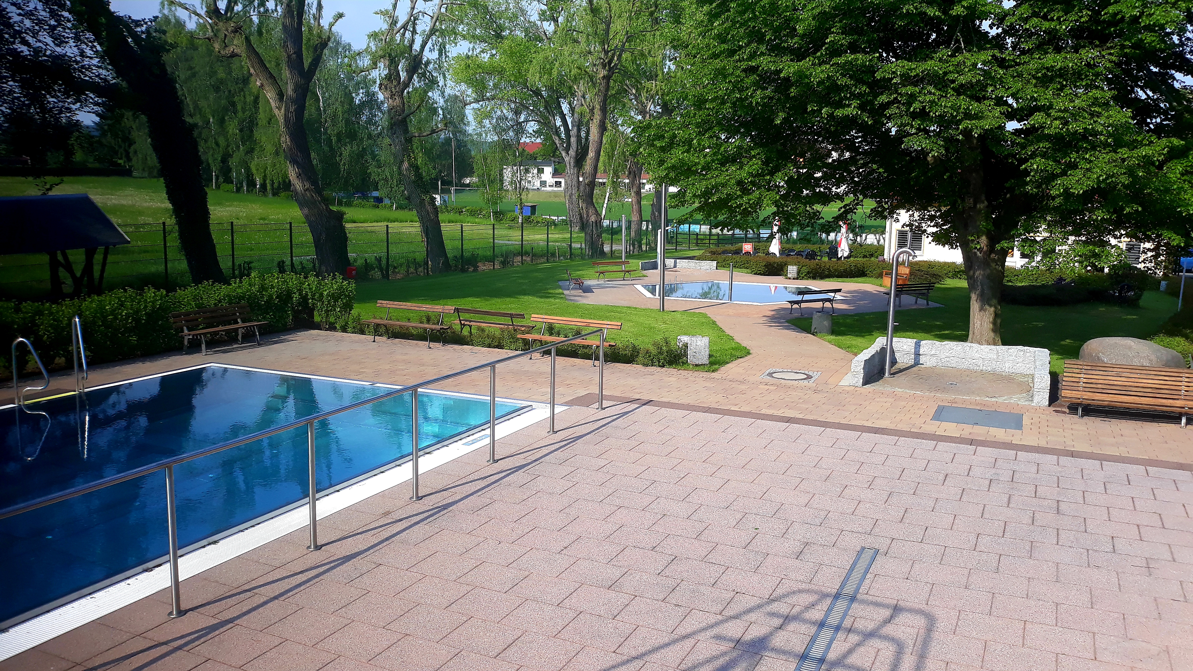 Das Bild zeigt Grünanlagen, im Vordergrund ist gepflasterter Untergrund und am linken Bildrand der Einstieg in ein Schwimmbecken zu sehen.