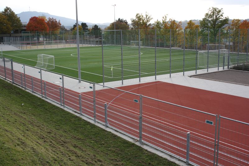 Das Bild zeigt einen Sportplatz mit Fußballfeld und roter Laufbahn sowie eine Kugelstoßanlage zu sehen. Die Felder umgibt ein hoher Zaun.
