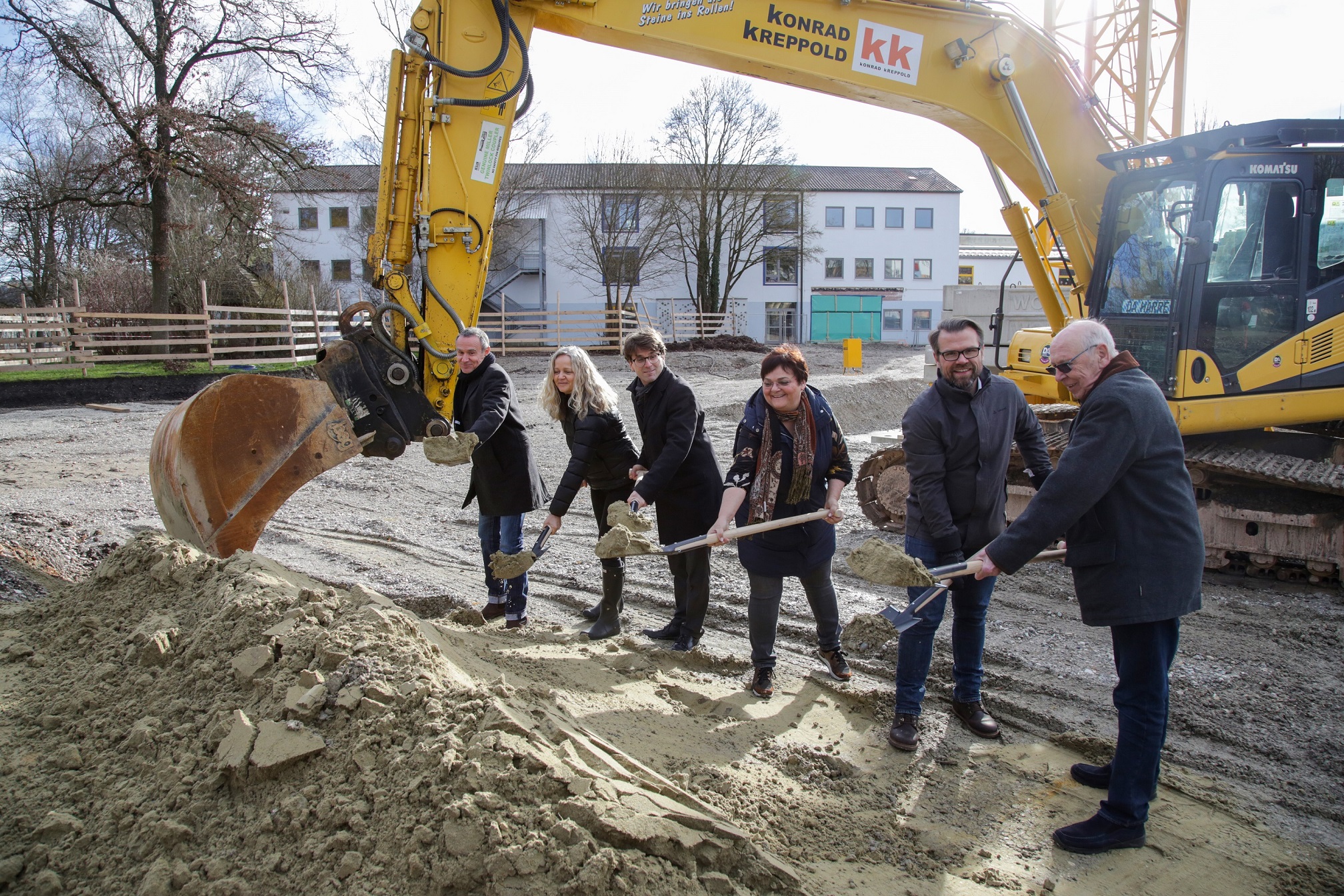Sechs Personen stehen hinter einem Sandhaufen und halten einen Spaten in der Hand, was den Baubeginn symbolisieren soll. Hinter den Personen stehet ein gelber Bagger.