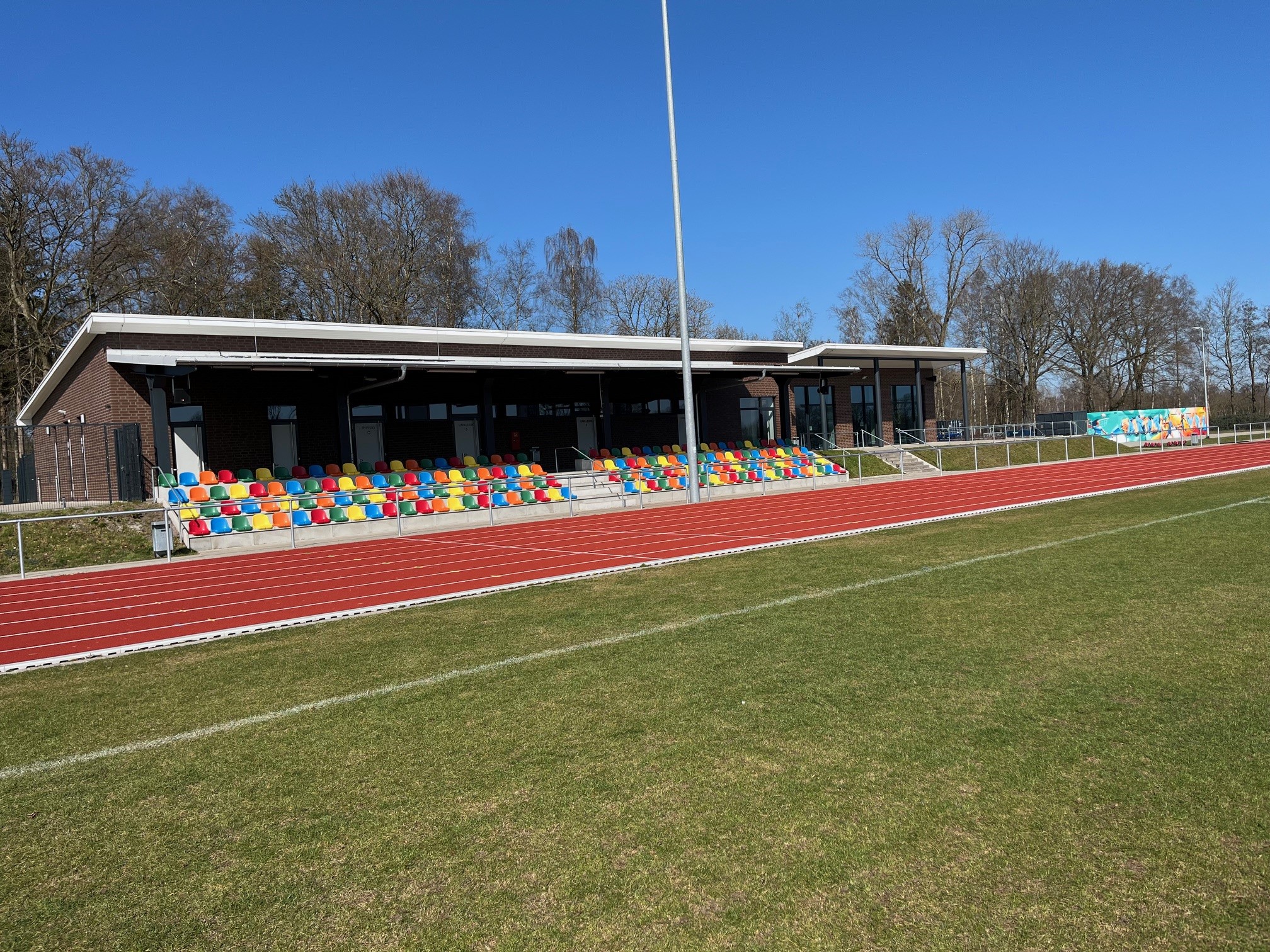 Stadt Varel Zu sehen ist eine Tribüne mit bunten Sitzschalen am Sportplatz, davor Laufbahn und Rasenfläche, im Hintergrund Bäume und blauer Himmel.