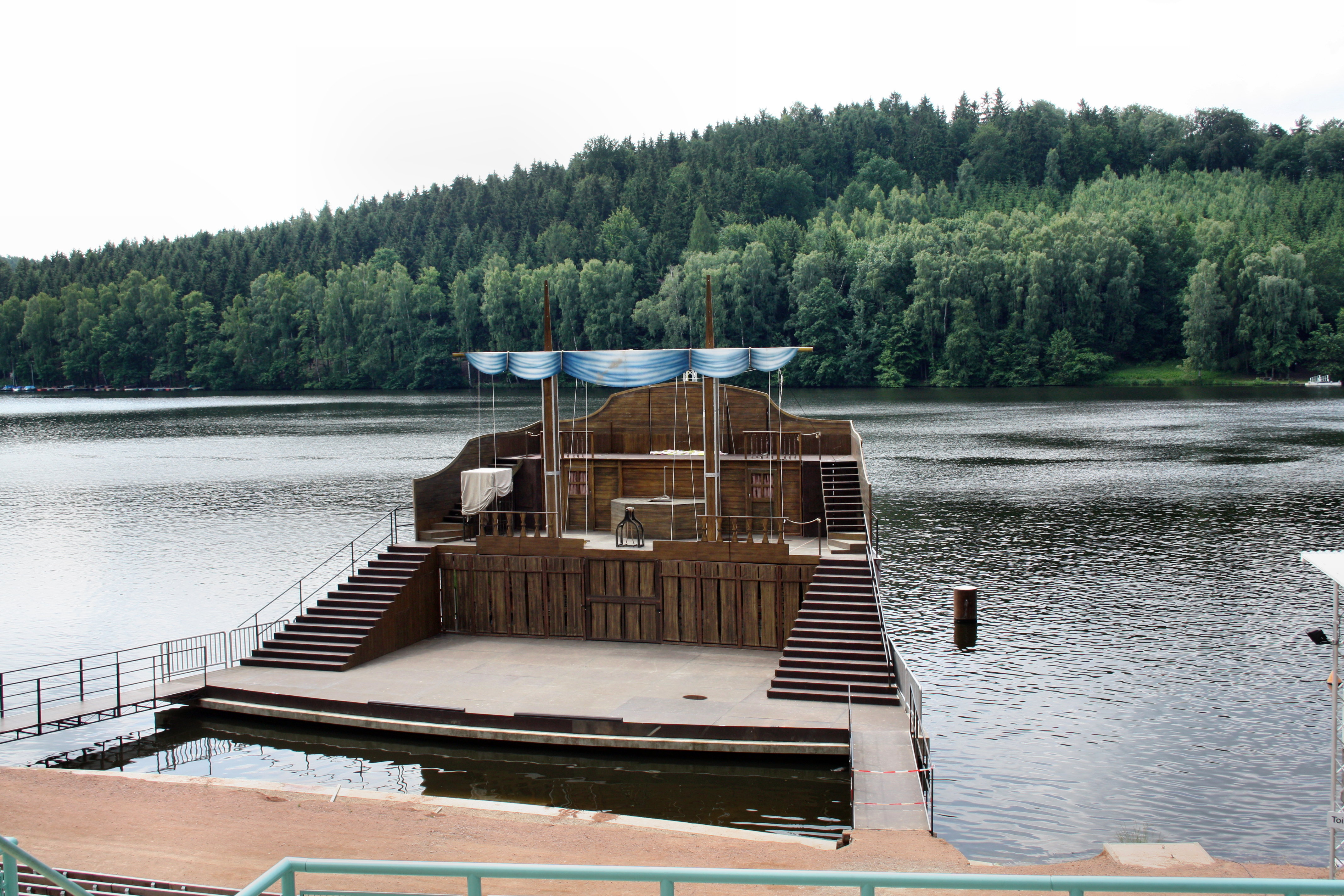 Das Bild zeigt eine Bühne aus Holz auf einem See. Im Hintergrund ist Wald.
