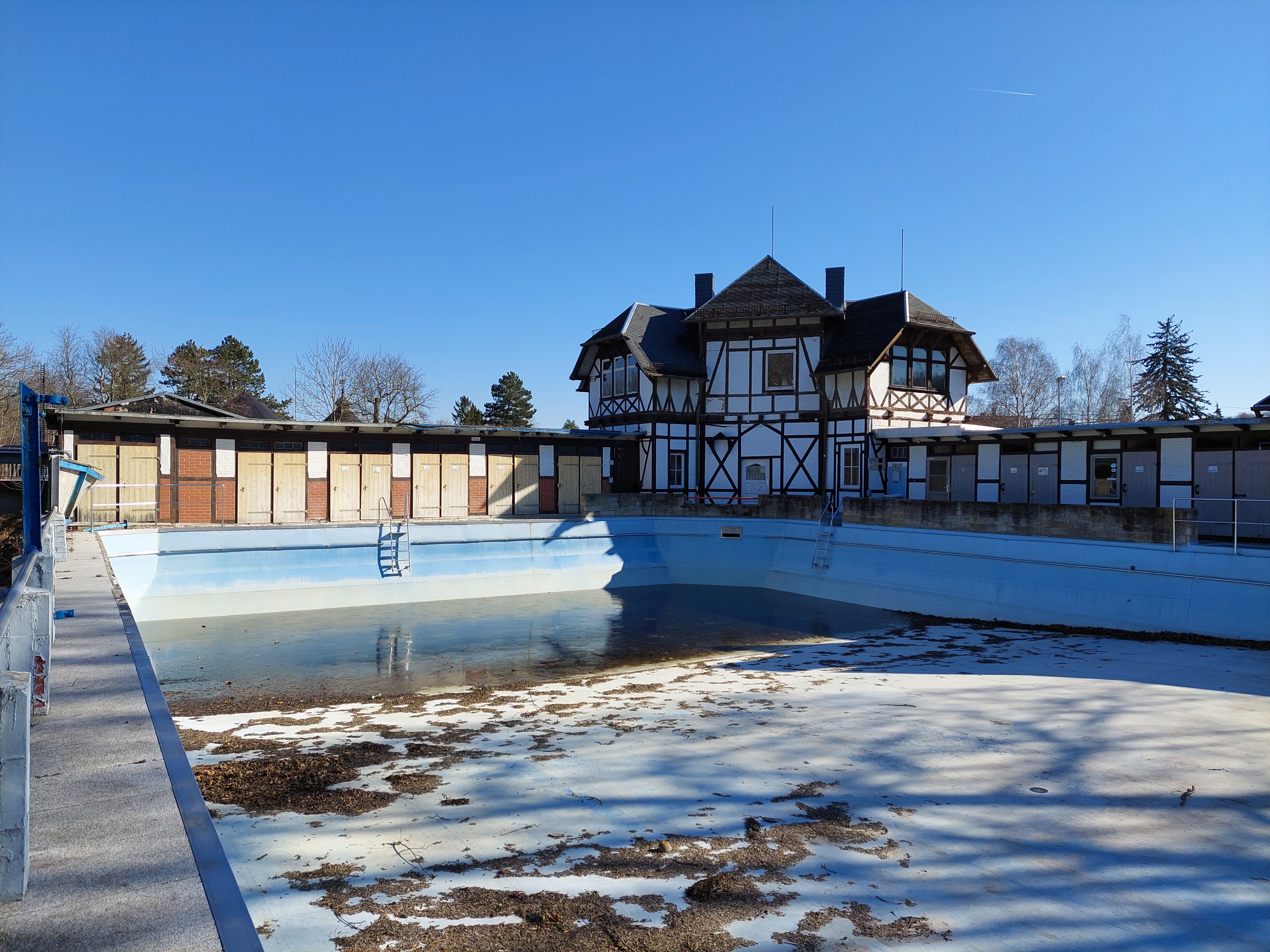 Das Bild zeigt ein Freibad im Winter. Das Becken ist leer. Im Hintergrund steht ein Fachwerkhaus.