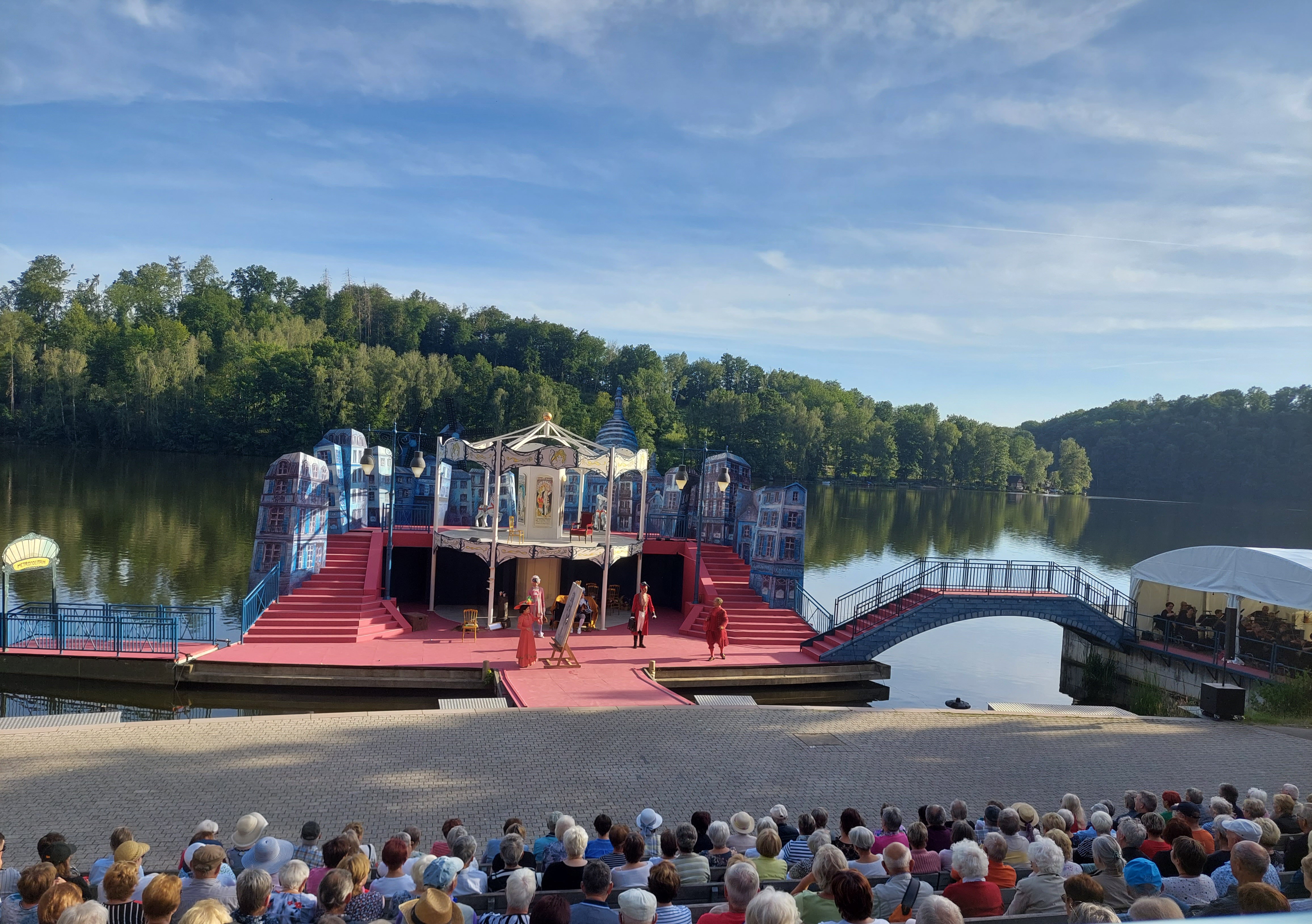 Das Bild zeigt die Bühne in Aktion. Die Treppe und Bühne sind mit rotem Teppich überzogen. Mehrere Schauspieler führen ein Stück auf. Im Vordergrund sitzen einige Personen auf der Zuschauertribüne. Im Hintergrund ist ein See und Wald zu sehen.