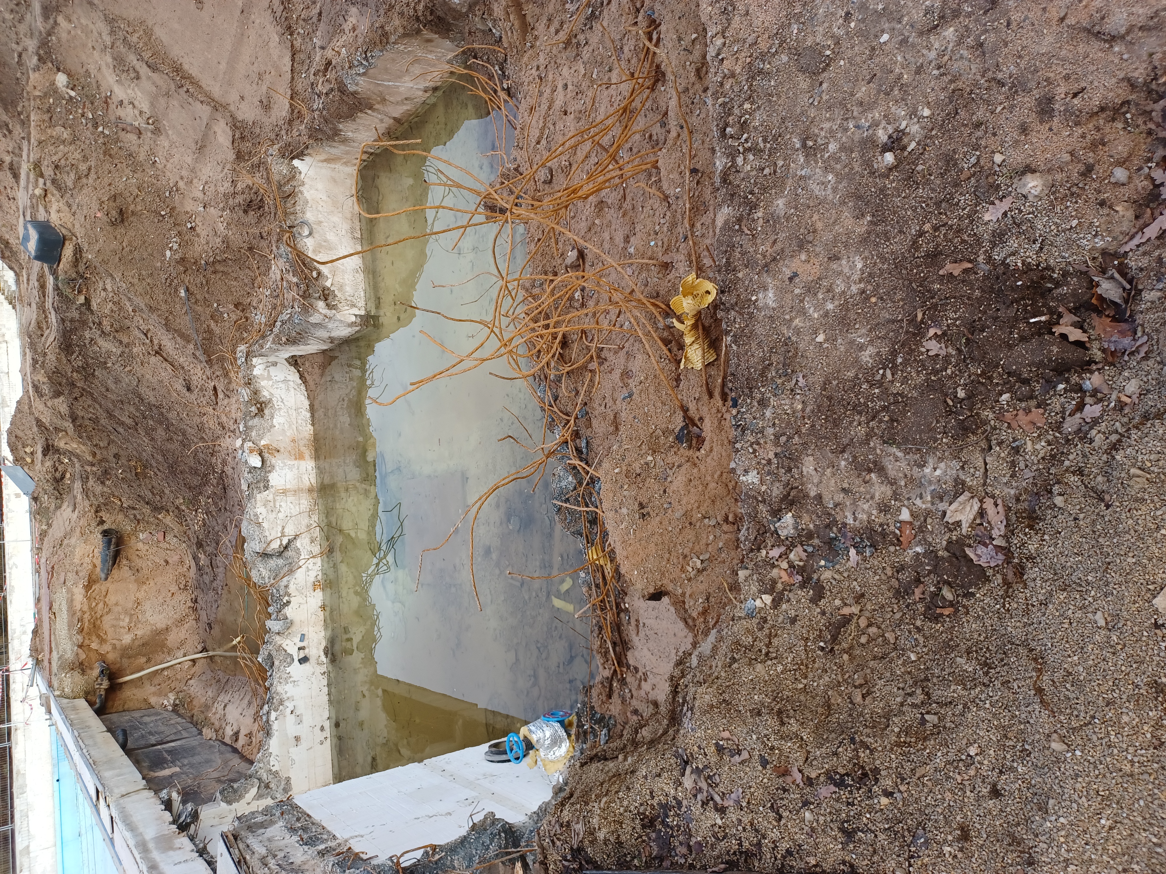Das Bild zeigt ein mit Wasser gefülltes Becken auf einer Baustelle. Aus dem Boden ragen Metallstangen. Der Rand ist von Erdhaufen umsäumt.