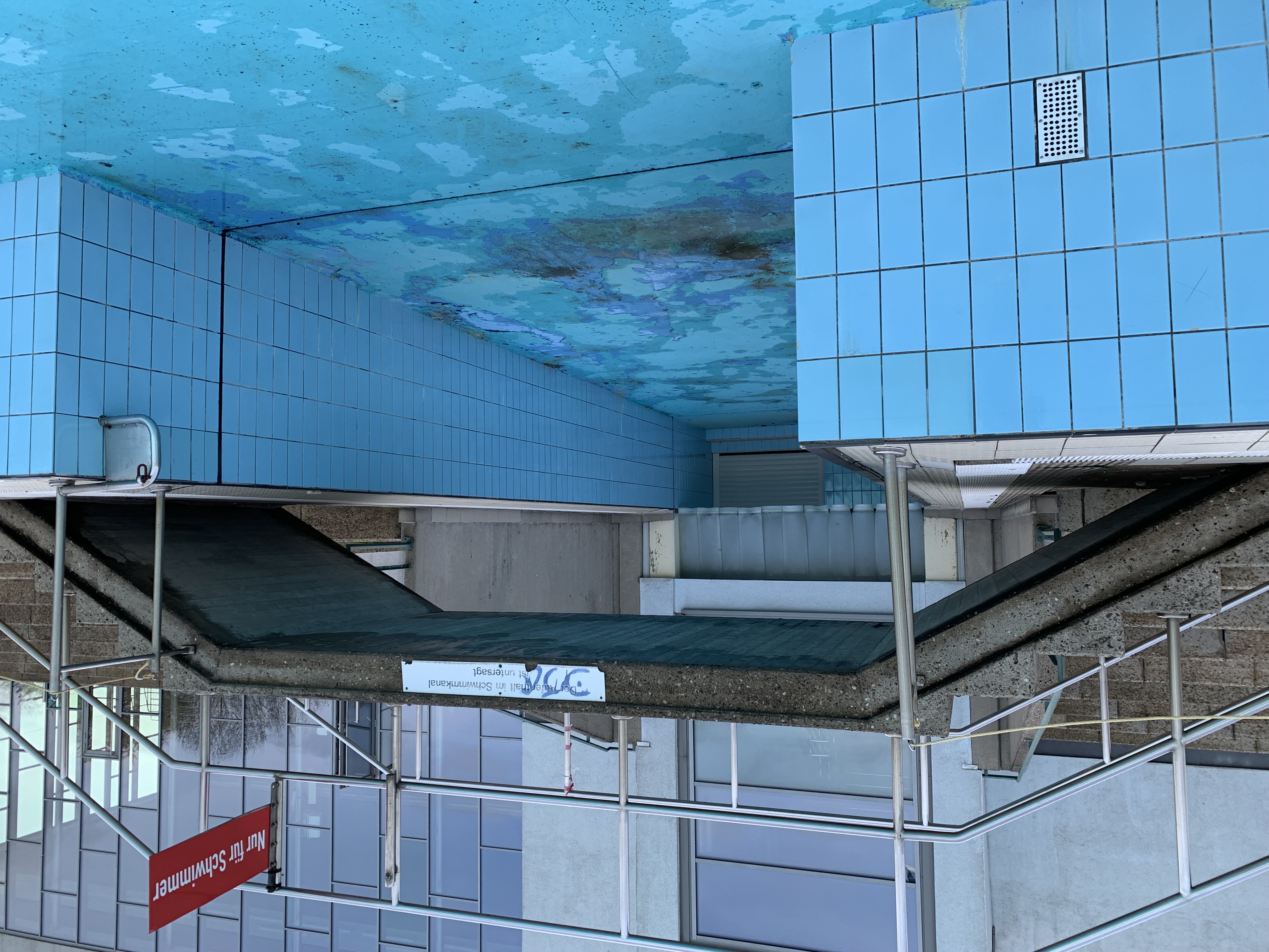 Das Bild zeigt ein leeres Schwimmbecken in einem Freibad. Eine Brücke bildet einen Fußweg über das Becken.
