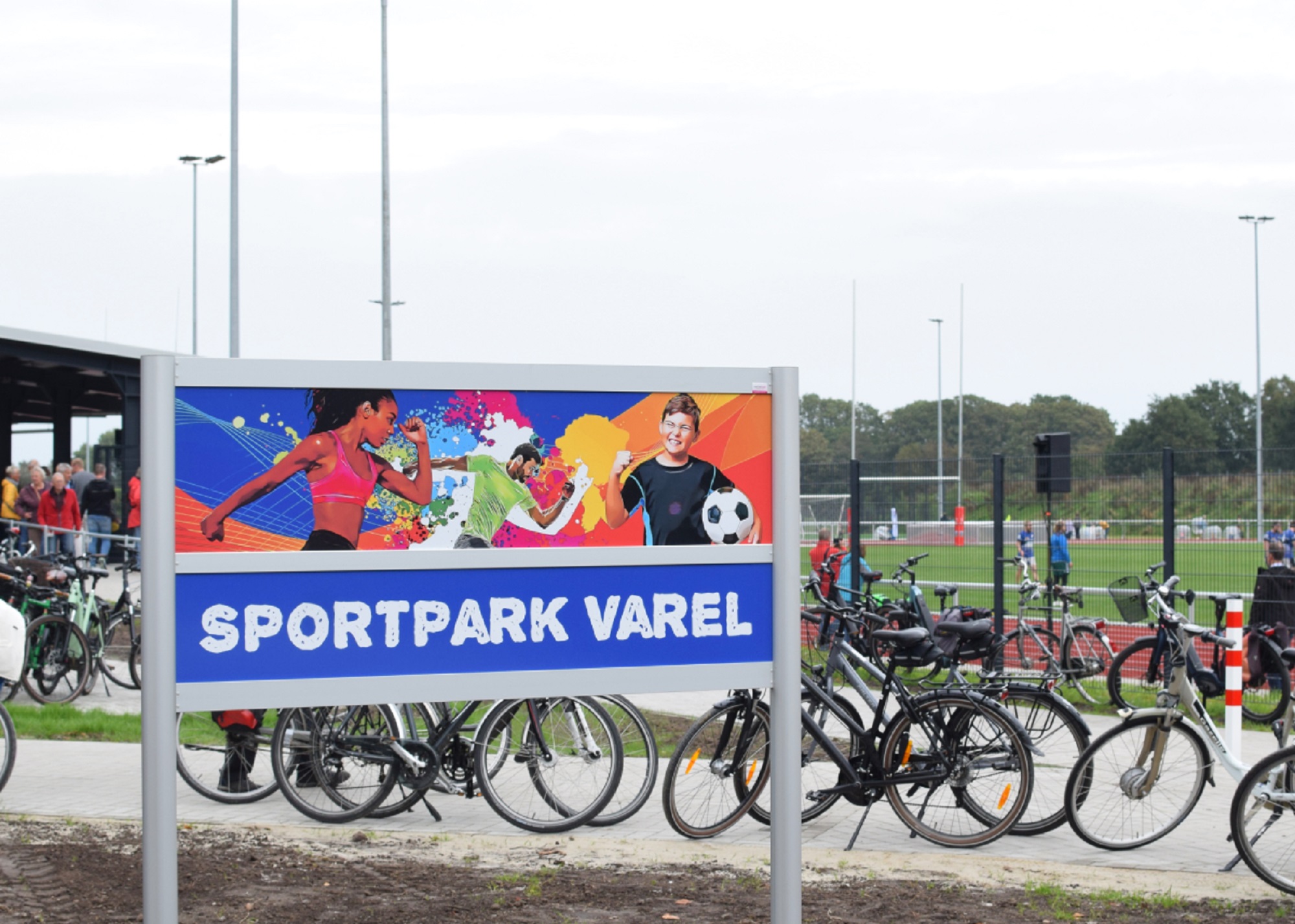 Stadt Varel Zu sehen sind ein Schild mit der Aufschrift 'SPORTPARK VAREL' vor Fahrradständern, im Hintergrund Sportplatz mit Spielern und Zuschauern.