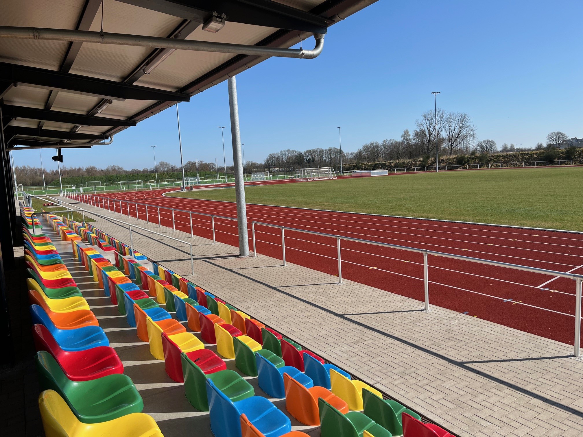 Stadt Varel Zu sehen sind bunte Sitzreihen unter einem überdachten Bereich neben einer roten Laufbahn und einem grünen Sportfeld im Sportpark.
